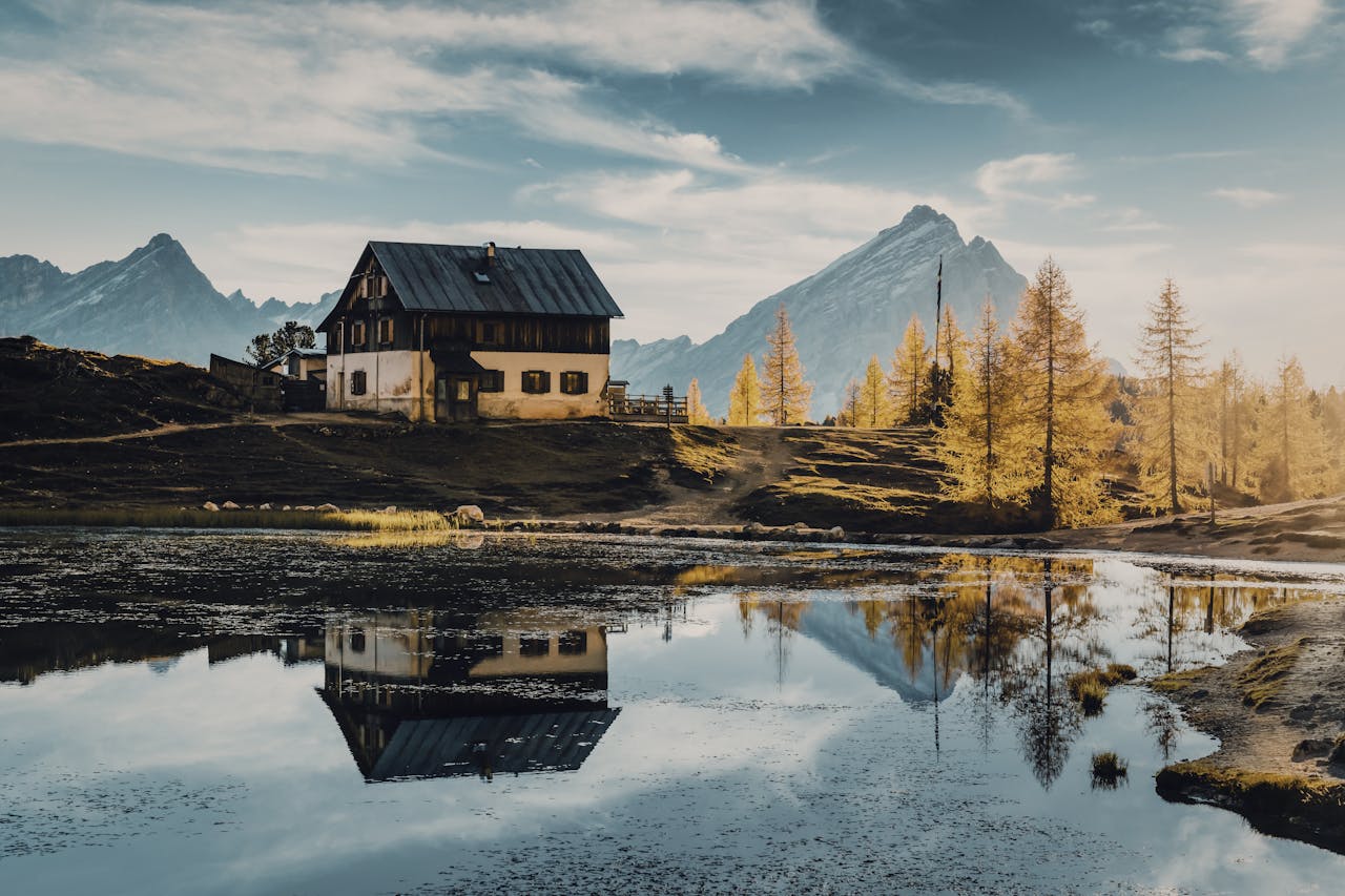 A rustic cabin and mountains reflected in a serene alpine lake under a clear sky.