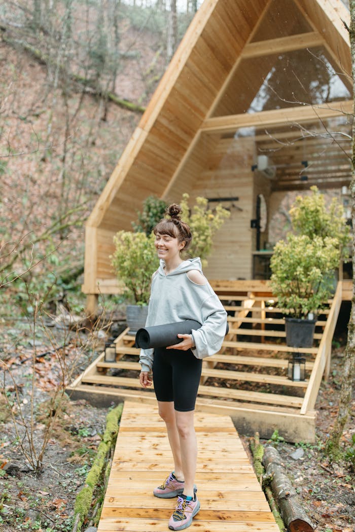 about-01 A woman enjoys a peaceful moment with a yoga mat in front of a modern wooden cabin in nature.